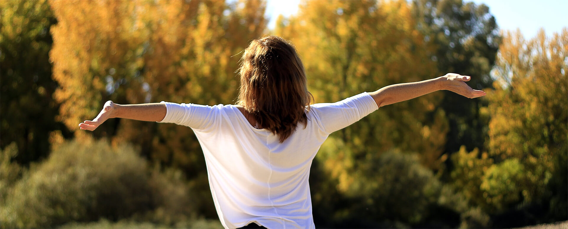 Eine junge Frau in einer weißen Bluse steht mit dem Rücken zu uns und hat die Arme ausgebreitet. Es ist ein sonniger Tag, im Hintergrund sieht man einen Herbstwald.