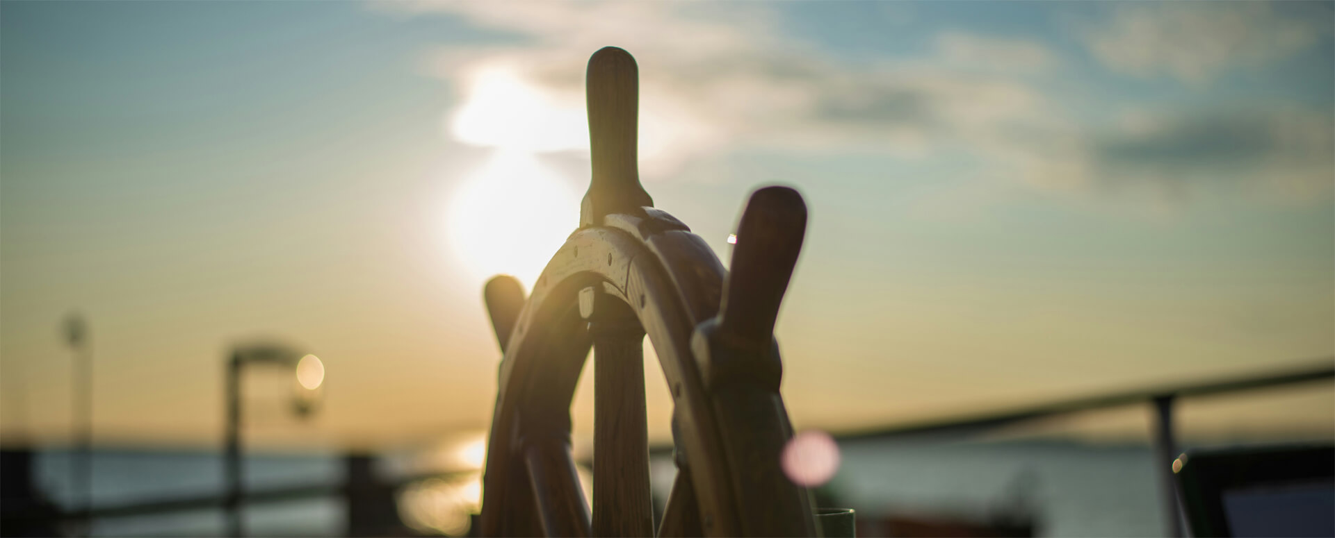 im Vordergrund sieht man ein hölzernes Steuerrad, im Hintergrund blauer Himmel, die Sonne wird durch da Steuerrad verdeckt.